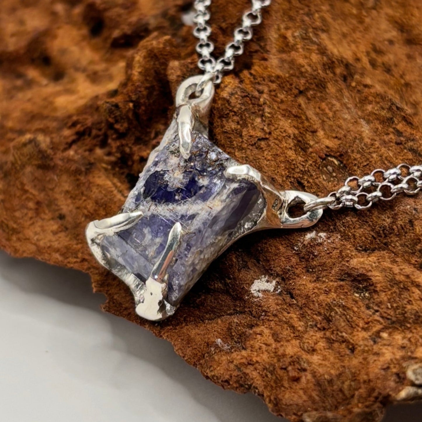 Silver pendant with a raw blue tanzanite stone on a chain, resting on a brown rock. handmade