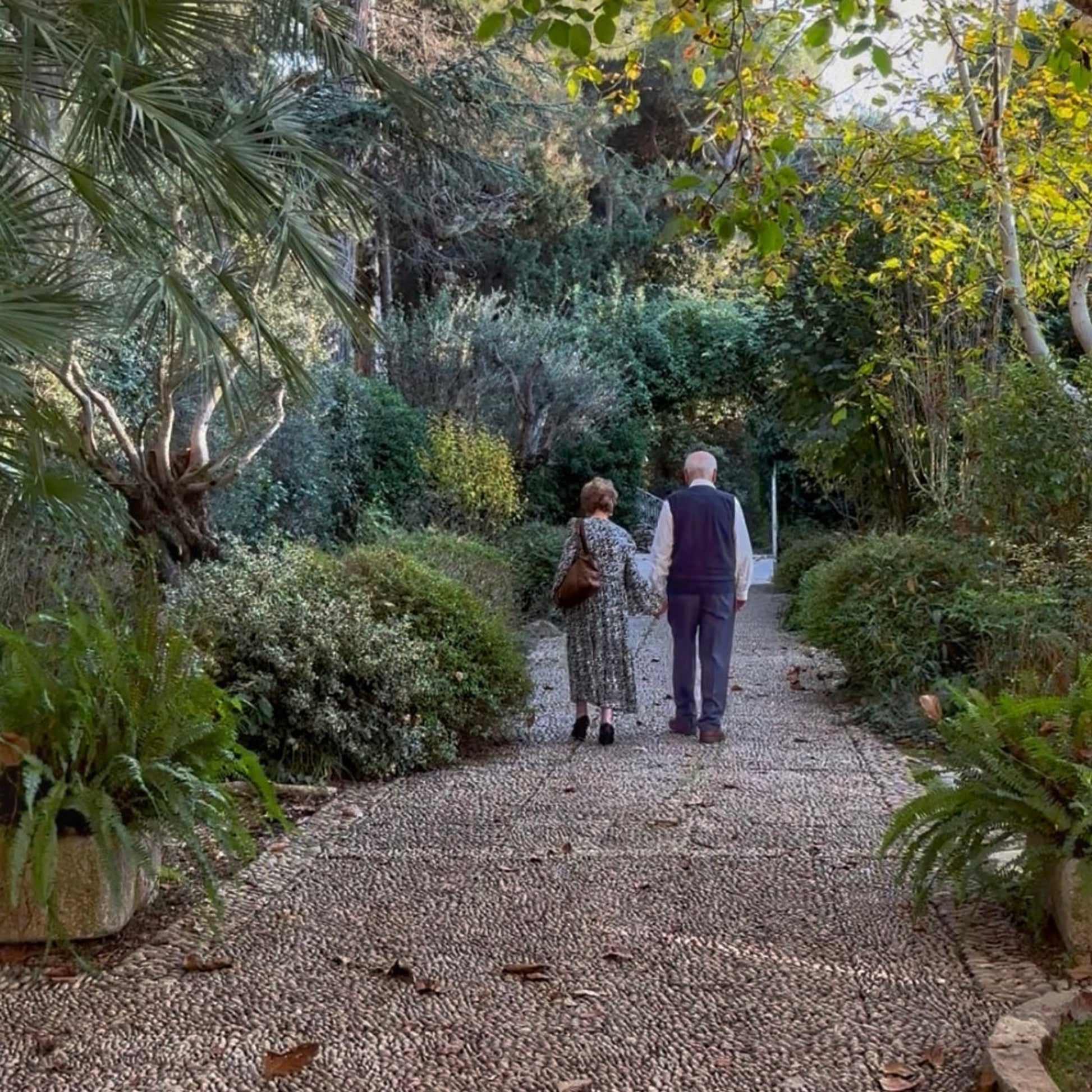 Two people walking along a gravel path in a garden with trees and plants.