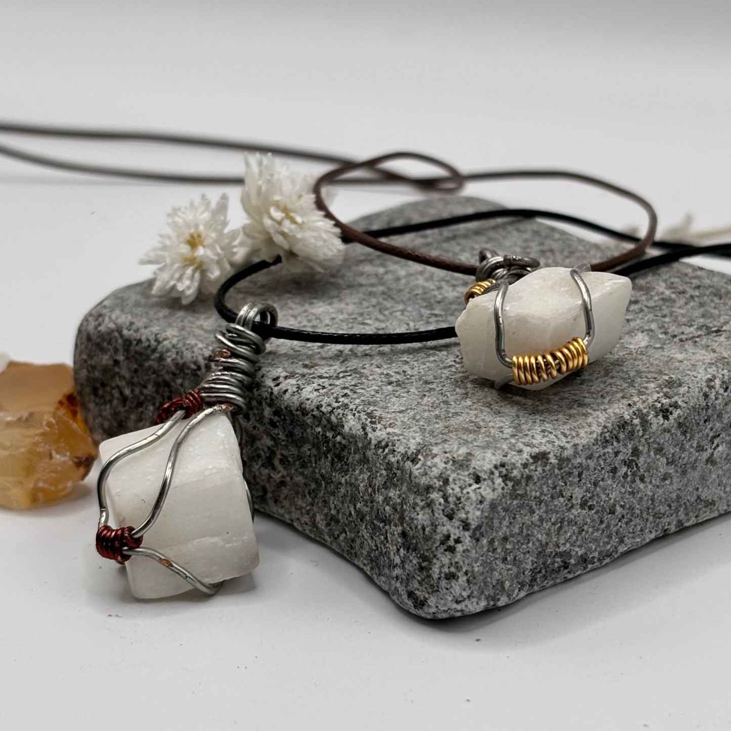 Two white cube stone pendants on leather cords with a white flower and crystal on a gray stone block.
