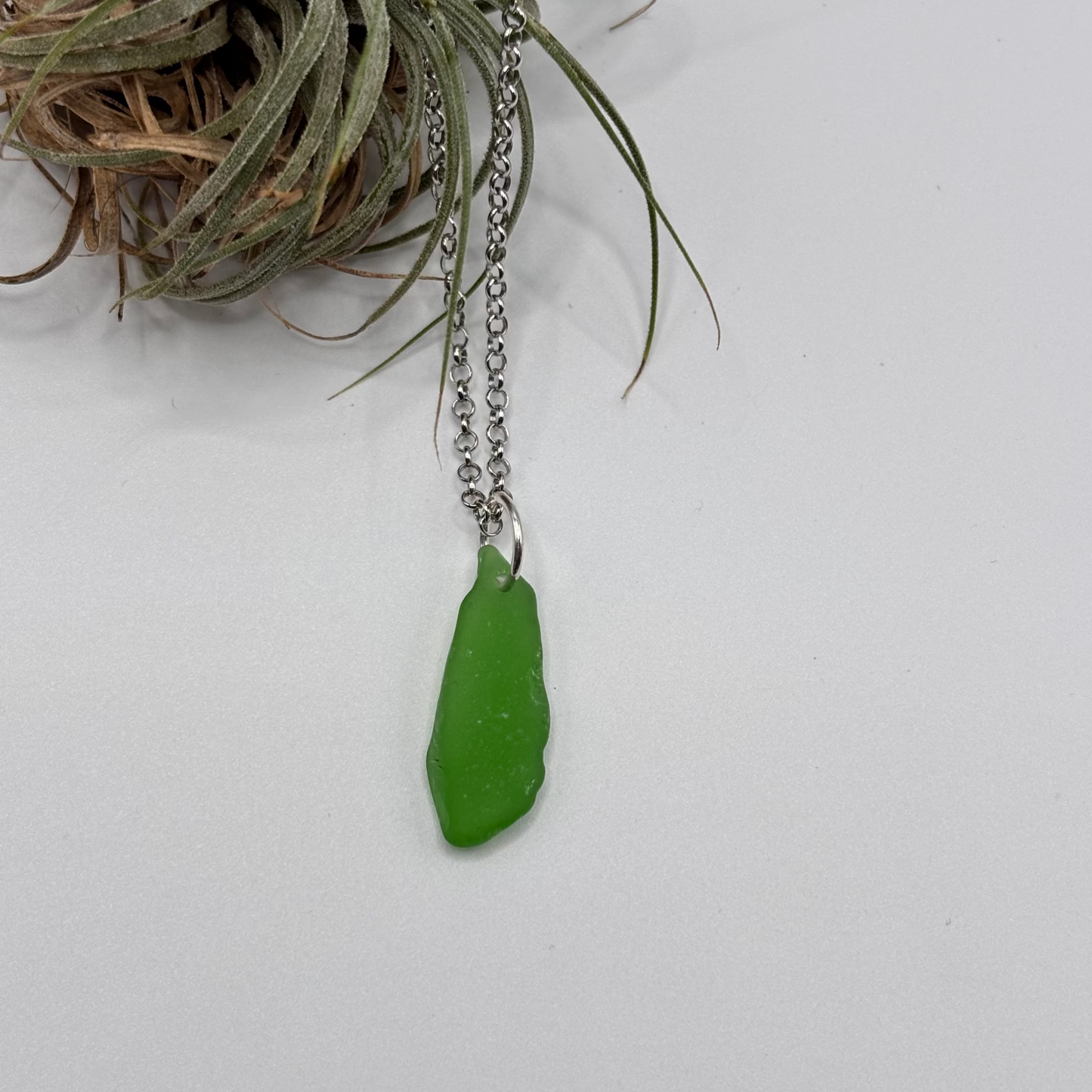 Green sea glass pendant with raw, tide-shaped edges, set in silver and hanging on a silver chain, capturing the natural movement and translucent glow of the sea.
