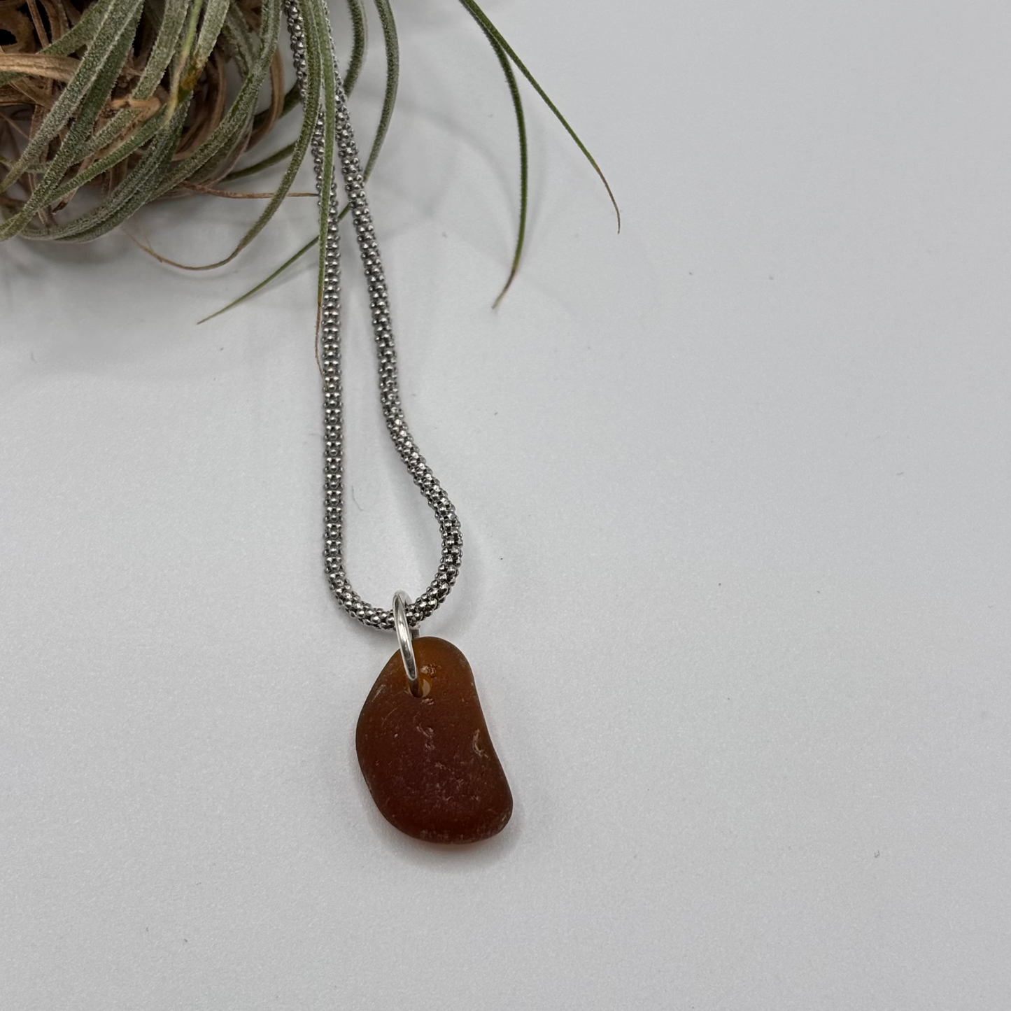 Brown sea glass pendant set in silver, suspended on a simple chain, photographed in soft natural light.