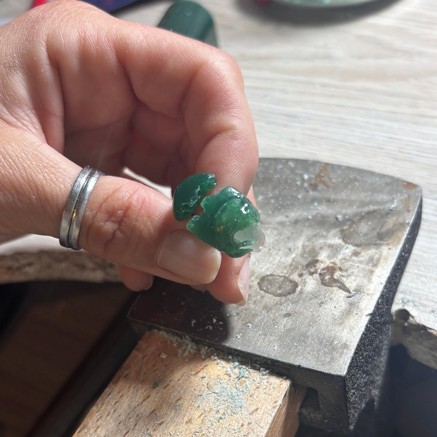 Hand holding a green wax model of a ring above a metal anvil on a workshop surface.