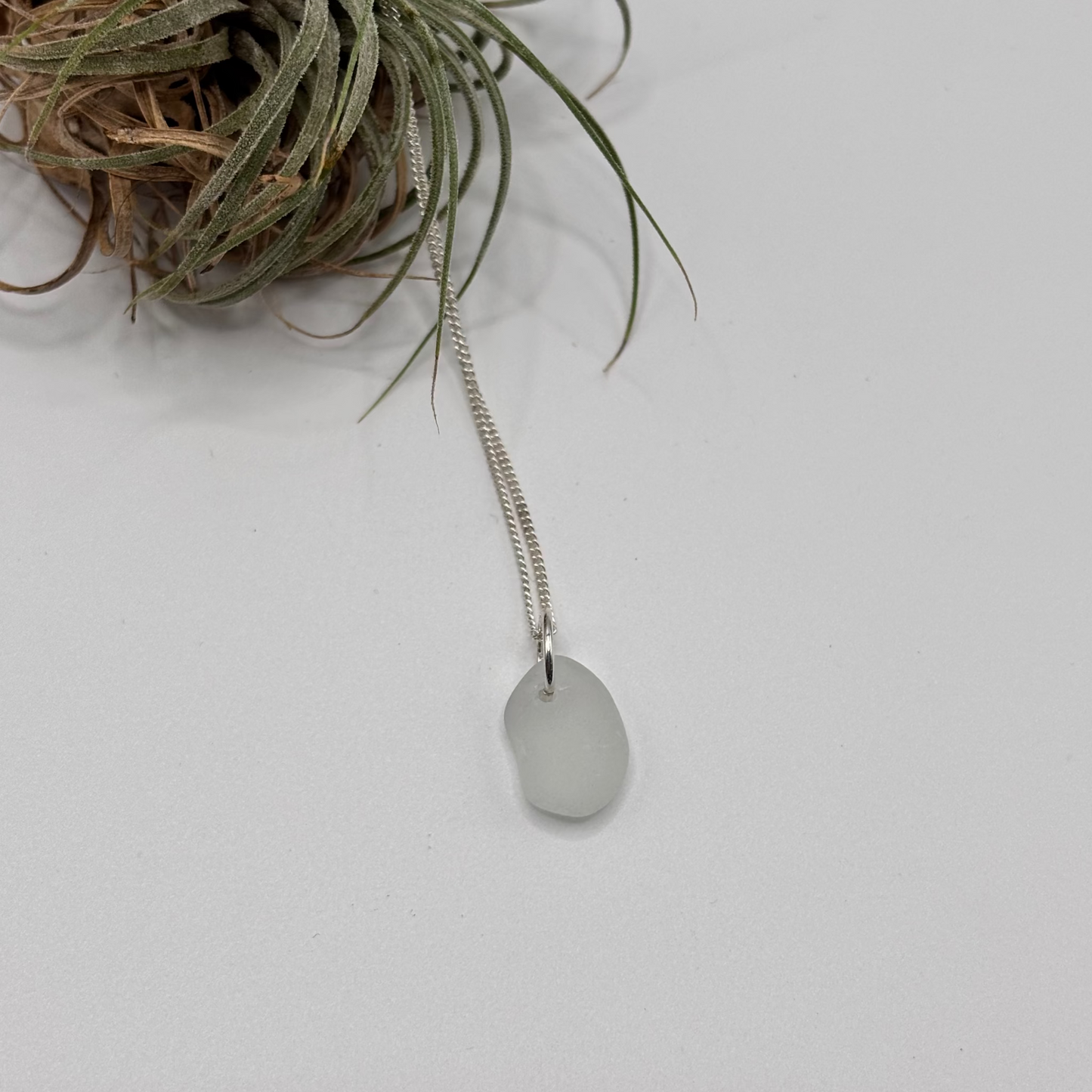 white small sea glass pendant with raw, tide-shaped edges, set in silver and hanging on a silver chain, capturing the natural movement and translucent glow of the sea foam.
