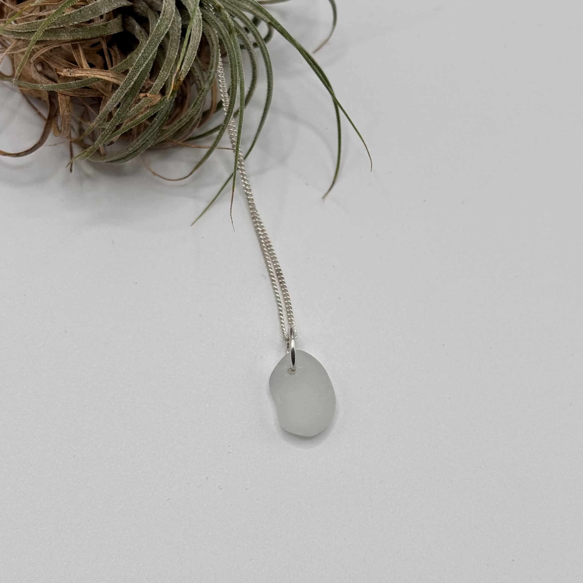 white small sea glass pendant with raw, tide-shaped edges, set in silver and hanging on a silver chain, capturing the natural movement and translucent glow of the sea foam.

